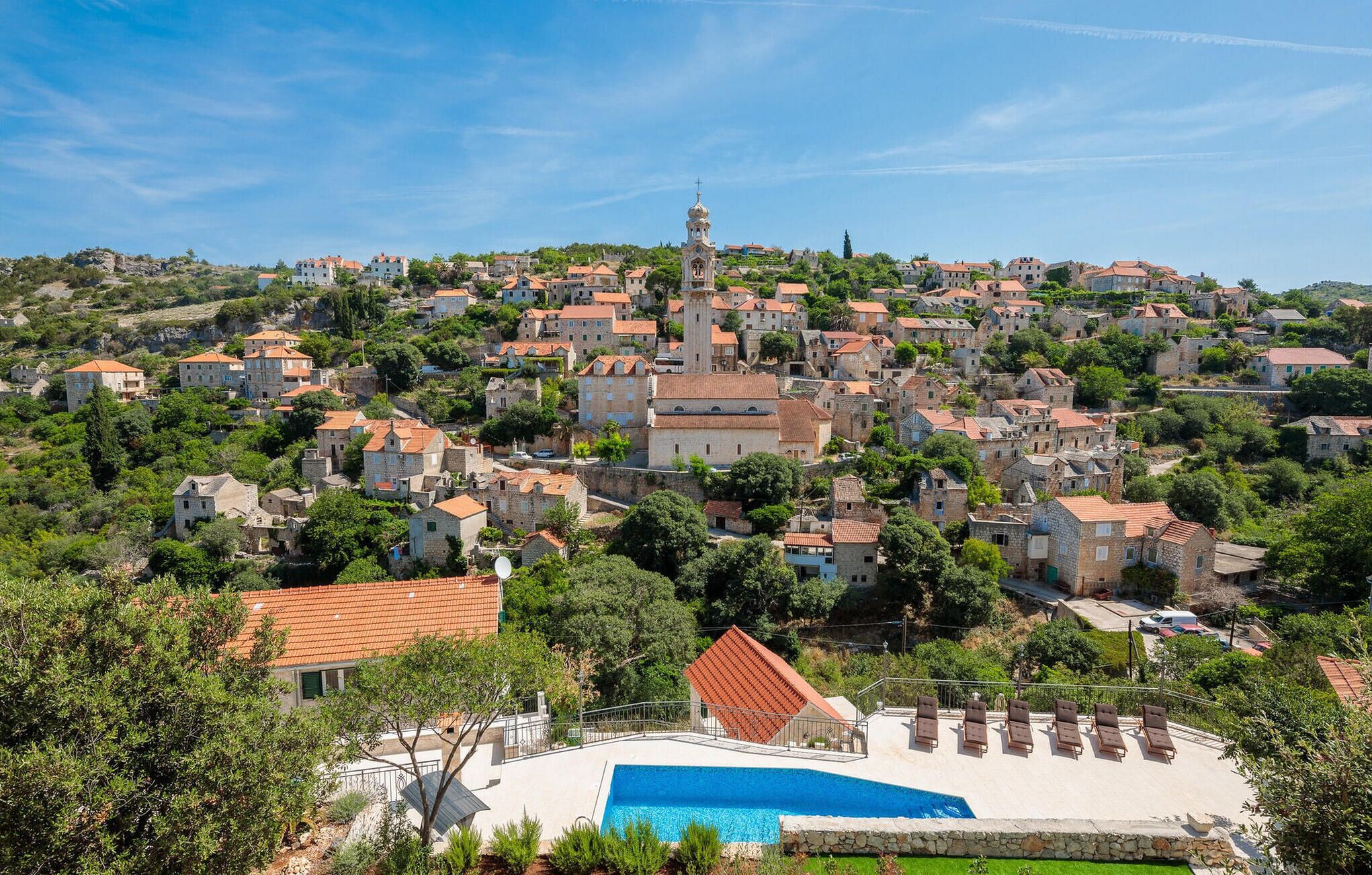 A hillside village with stone buildings, a prominent church, and a swimming pool area.