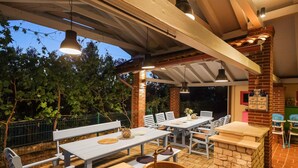 An outdoor patio with multiple tables and benches, a stone fireplace, and a covered area with hanging lights.