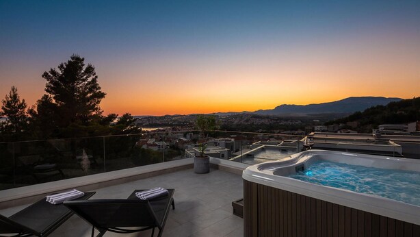 A rooftop terrace with a hot tub, lounge chairs, and a view of a cityscape at sunset.