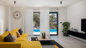 A modern living room with a yellow sofa, a coffee table with a pineapple centerpiece, a large clock, and a view of a swimming pool through the glass door.