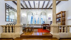 Una habitación amplia con suelo de madera, alfombra roja, escritorio, silla, estantería y ventanas grandes con cortinas.