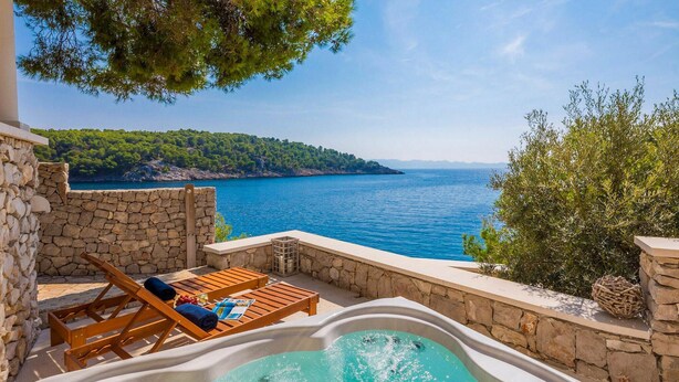 Un espacio al aire libre con paredes de piedra, un jacuzzi, sillones de madera y vista al mar.