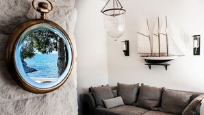 Una habitación con un espejo redondo que refleja un árbol y agua, un sofá con cojines, una lámpara colgante y un modelo de velero sujeto a la pared.