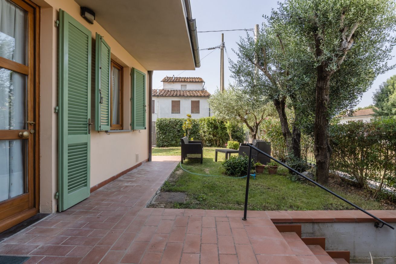 A house with a brick pathway leading to a door, green shutters, and a garden with a table and chairs.