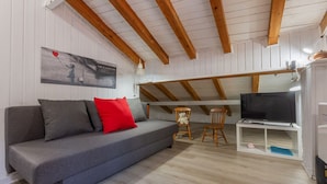 Une pièce chaleureuse avec un canapé gris, des coussins rouges et gris, un téléviseur posé sur un meuble blanc et un plafond en bois.