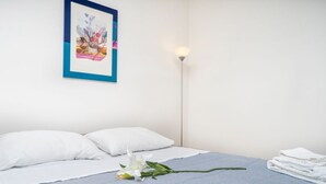 Un lit bien fait, avec des oreillers blancs et une couverture bleue, une seule fleur blanche posée sur le lit, une œuvre d’art encadrée accrochée au mur et une lampe sur pied à côté du lit.