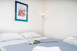 A neatly made bed with white pillows and a blue blanket, a single white flower on the bed, a framed artwork on the wall, and a floor lamp beside the bed. - GuestHost - A Cozy Nest in the City - Studio for 2! (Udine)