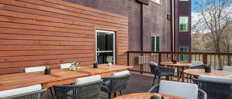An outdoor patio with wooden tables and chairs, a view of a building with large windows, and a clear sky.