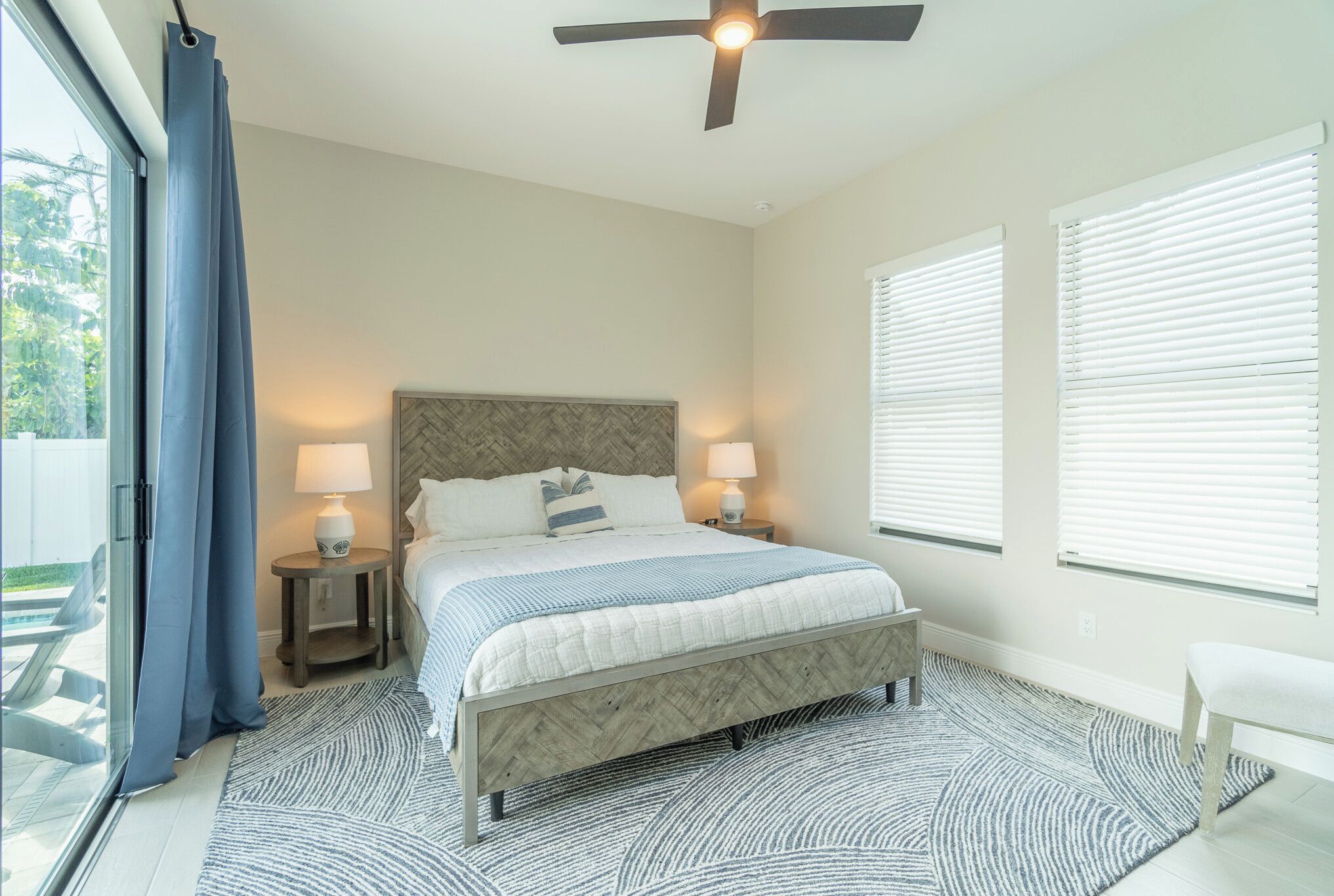 A bedroom with a bed, two bedside lamps, a ceiling fan, and a view of the outdoors.