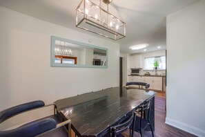 Een eetgedeelte met een lange tafel, stoelen, een kroonluchter, een spiegel en een keuken op de achtergrond.