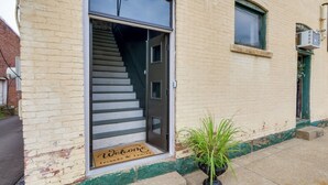 Entrada de edificio con felpudo de madera, planta en maceta y una escalera que conduce al interior.