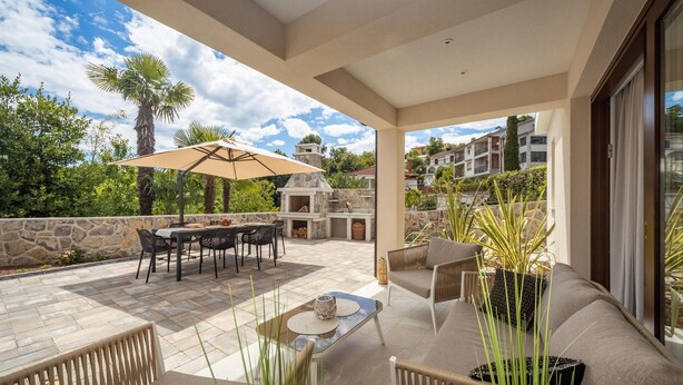 Une terrasse extérieure couverte, dot ée d’un sol en pierre, d’une table à manger avec des chaises, d’un canapé et d’un grand parasol.