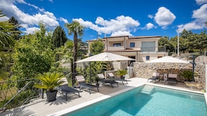Un espace aménagé au bord de la piscine, avec des chaises longues, un mur en pierre et un bâtiment à deux étages en arrière-plan.