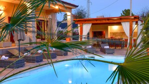 A swimming pool with lounge chairs and umbrellas, a covered outdoor seating area, and a stone facade building.