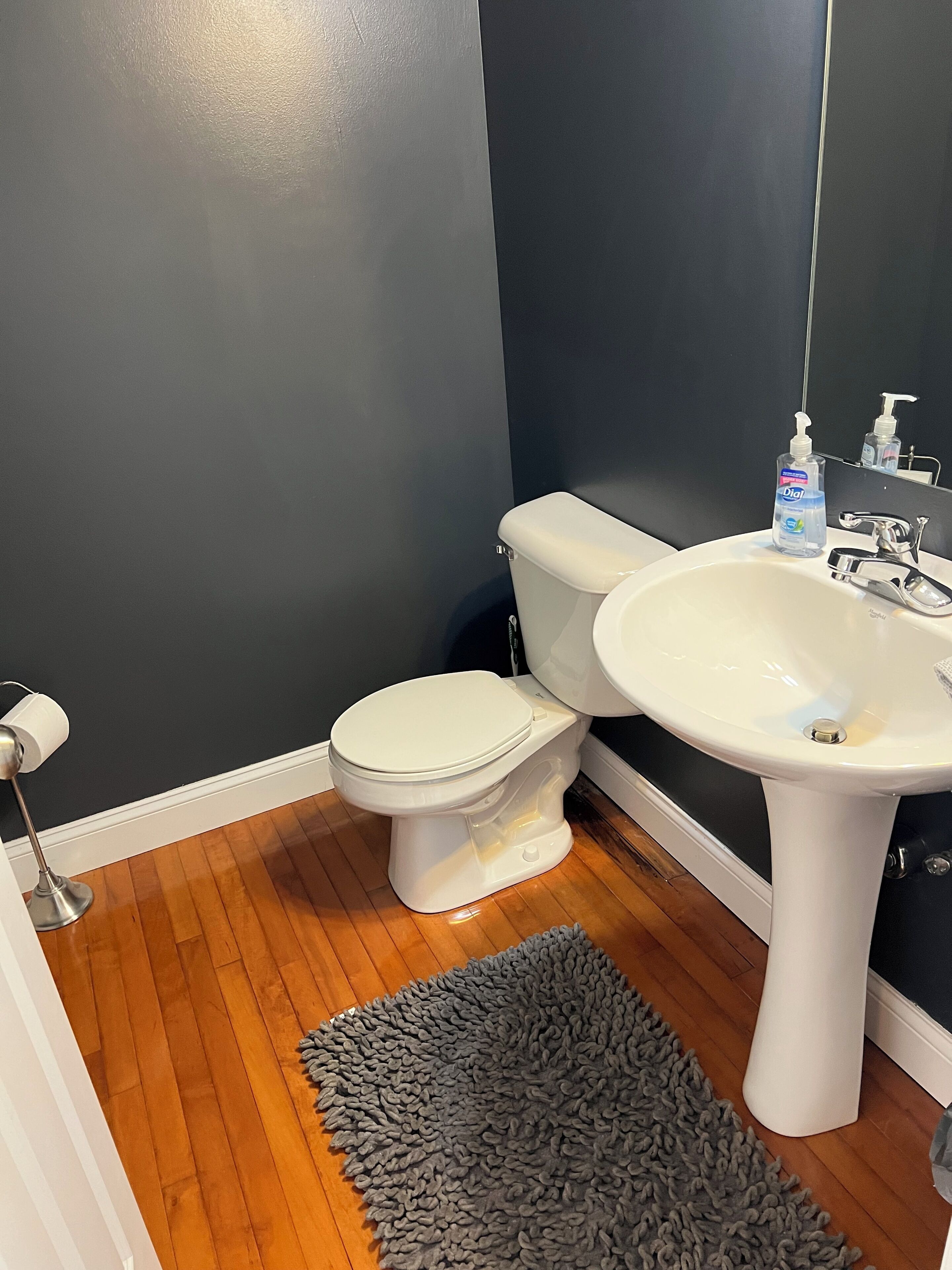 Un baño con inodoro y lavamanos blancos, pared oscura y suelo de madera.