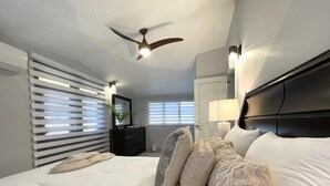 A modern hotel room with a bed, a ceiling fan, a nightstand, a lamp, and a window with blinds.