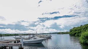 A marina with boats docked at wooden piers, surrounded by lush greenery and a calm lake under a partly cloudy sky.