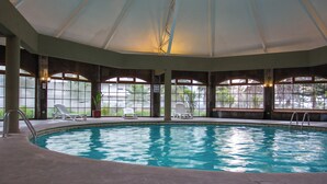 Una piscina interior con paredes de vidrio transparente, suelo de baldosas y varias sillas de descanso.