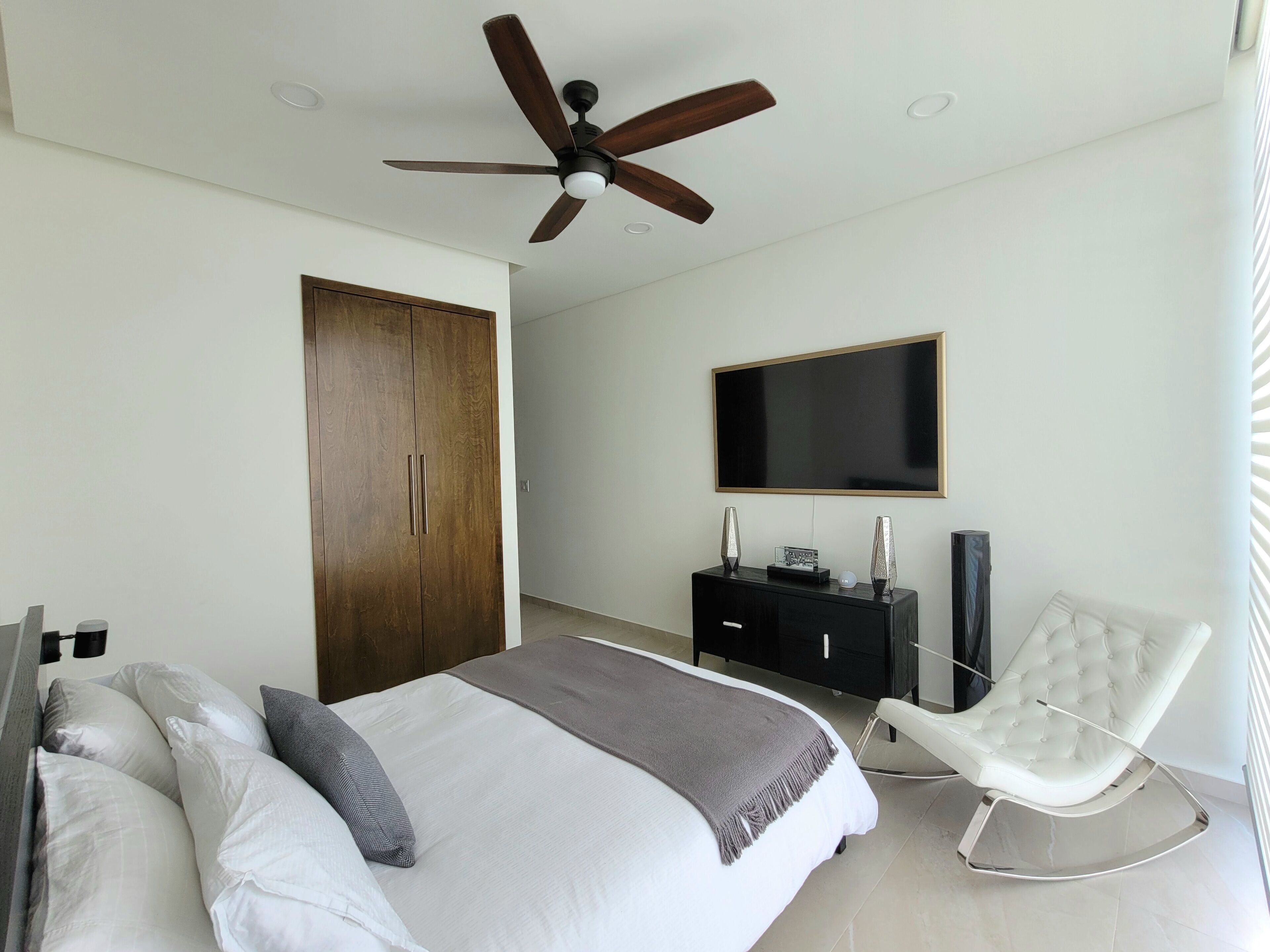 Un dormitorio con cama, televisor, armario, silla y ventilador de techo.