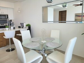 A modern kitchen with white cabinets, a microwave, and a coffee machine. There is a round glass table with white chairs and a decorative metal piece on it. - Luxury Beachfront Condo in Rosarito with Pool & Jacuzzi (Playas de Rosarito)