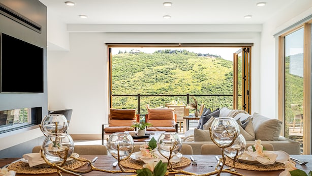 Un coin repas avec une table dressée pour quatre personnes, un canapé et une grande fenêtre donnant sur une colline verdoyante.