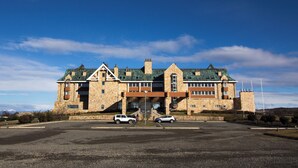 A large stone building with a green roof, two cars parked in front, and a clear blue sky. - Hotel y Cabañas Llanuras de Diana- Caja Los Andes (Natales)