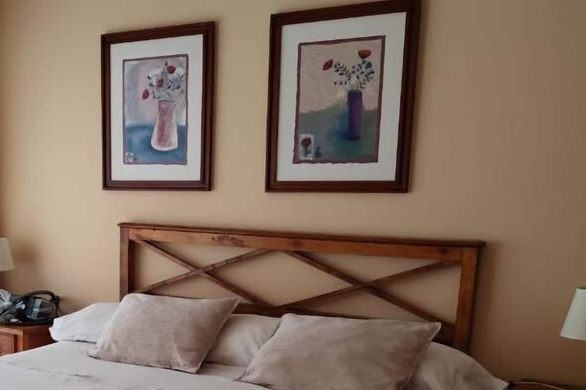 Ein ordentlich bezogenes Bett mit einem Holzkopfteil, ein Nachttisch mit einer Lampe sowie zwei gerahmte Kunstwerke an der Wand.