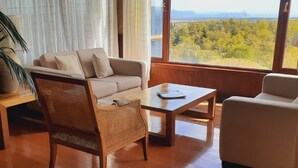 A room with a sofa, a wicker chair, a coffee table, and a view of a landscape through large windows. - Hotel y Cabañas Llanuras de Diana- Caja Los Andes (Natales)