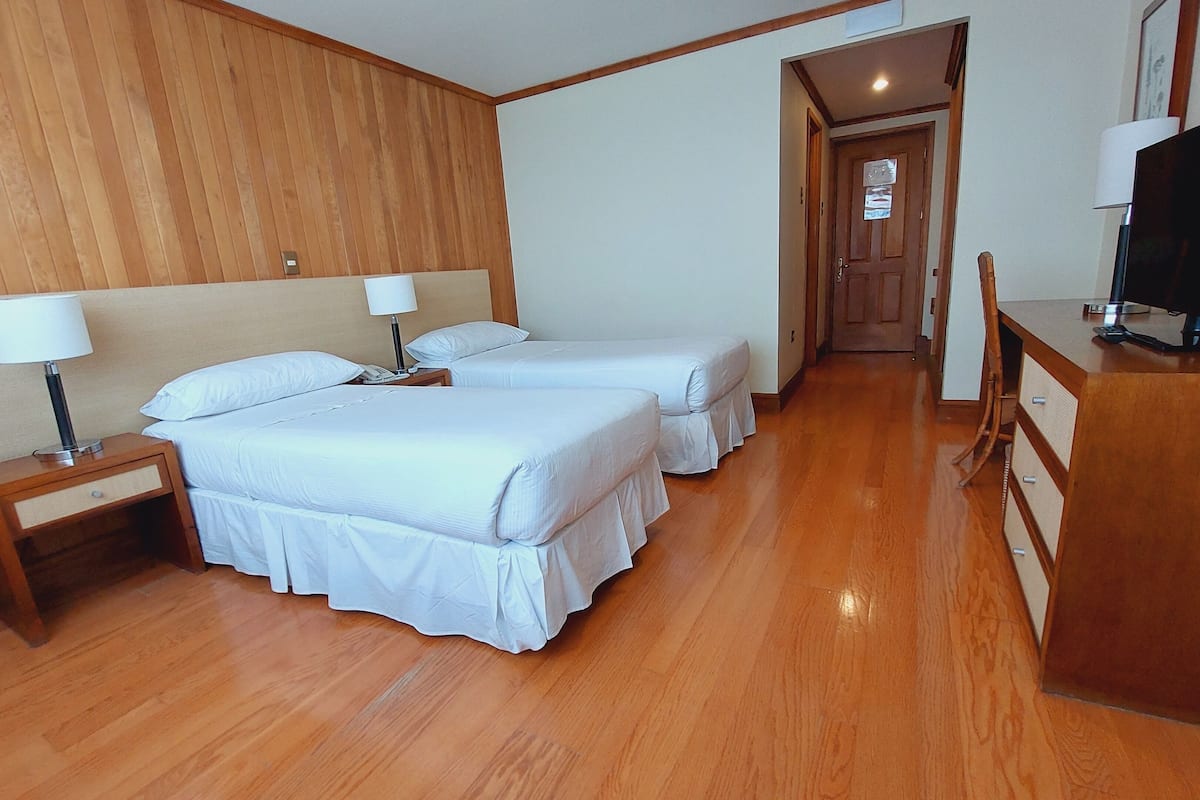 Ein Hotelzimmer mit zwei Betten, Holzboden, einem Schreibtisch mit Fernseher und einer Tür zu einem weiteren Raum.