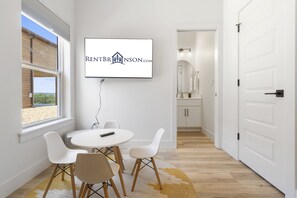 A modern living room with a TV mounted on the wall, a dining table with chairs, and a view of a sandy landscape through a window. - Private Hot Tub & Deck with Best Table Rock Views + $400 Free Tickets (Kimberling City)
