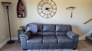 A living room with a dark brown leather sofa, a round wall clock, a fish on a wall, and a small shelf with various items. - Ozarks View with Cornhole, Pickleball, Community Pools, Firepit,  Free Tickets and More (Kimberling City)