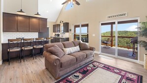 A spacious living area with a brown sofa, a dining table with chairs, a ceiling fan, and a view of a deck with a "WELCOME" sign. - Ozarks View with Cornhole, Pickleball, Community Pools, Firepit,  Free Tickets and More (Kimberling City)