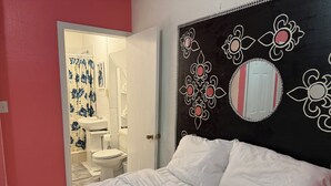 Een slaapkamer met een bed, een wit hoofdeinde met bloemmotieven, en een badkamer zichtbaar door een open deur.