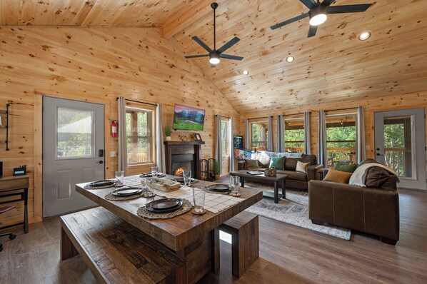 Una cabaña de madera espaciosa con comedor, chimenea, sofá y ventanas grandes.