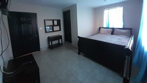 A bedroom with a bed, a small table, a mirror, a door, and a TV. - Departamentos Velasco 2 (Tijuana)