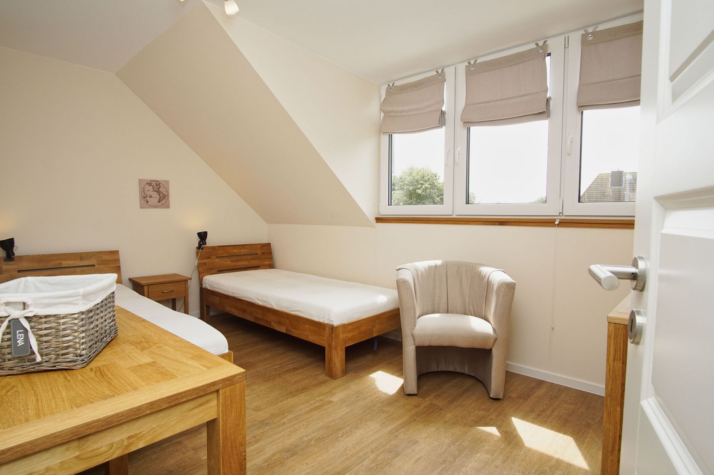 A small, well-lit room with a sloped ceiling, a bed, a chair, a table, and a window with curtains.