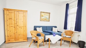 Ein Zimmer mit einem Holzschrank, einem blauen Sofa, zwei Korbstühlen, einem kleinen Tisch mit einer blauen Tasse und einem Fenster mit Vorhängen.