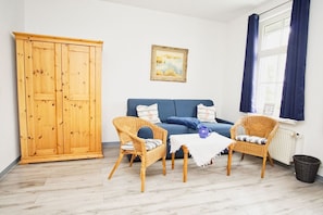 Ein Zimmer mit einem Holzschrank, einem blauen Sofa, zwei Korbstühlen, einem kleinen Tisch mit einer blauen Tasse und einem Fenster mit Vorhängen.