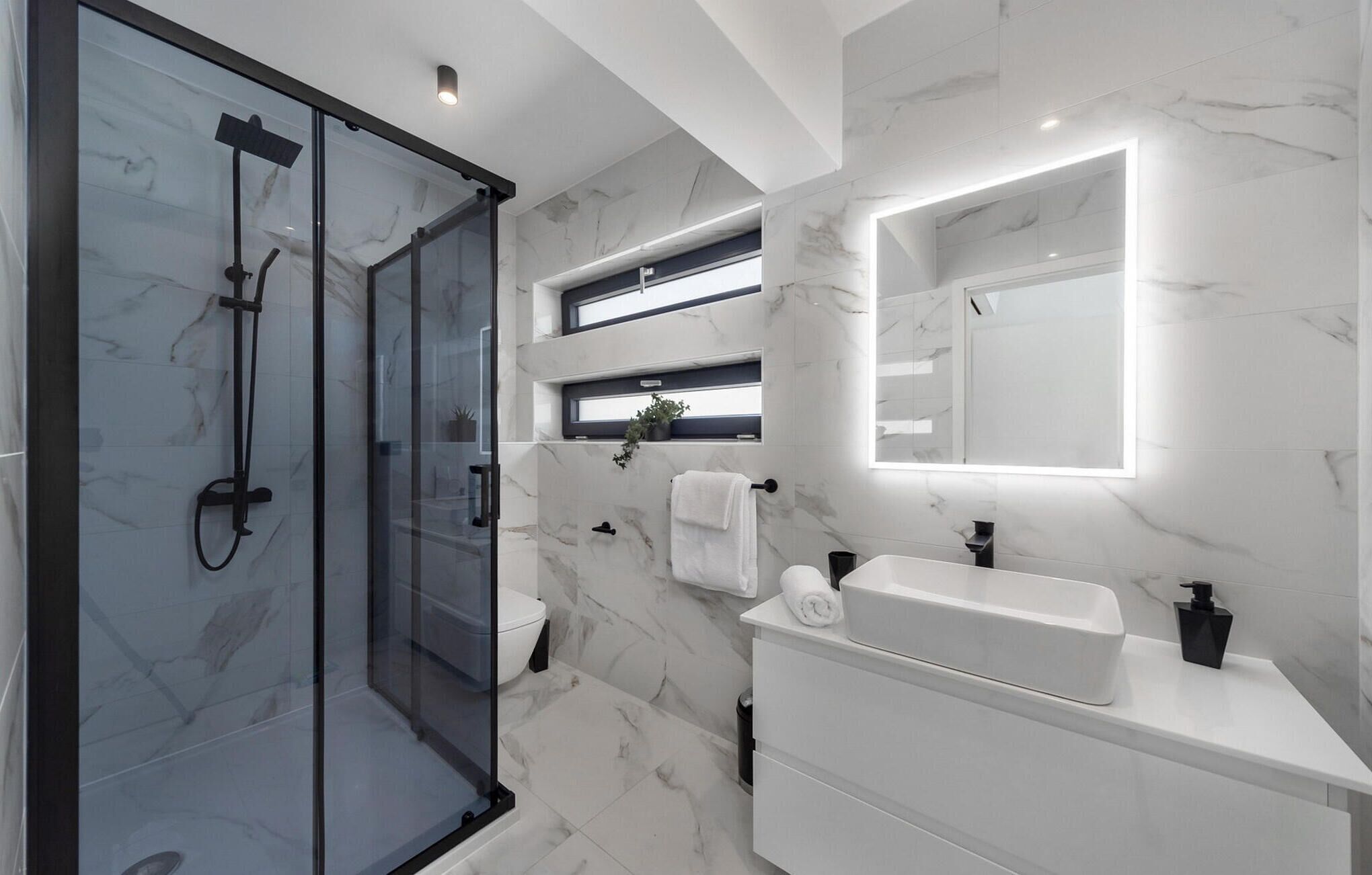 Een moderne badkamer met een glazen douchecabine, een witte wastafel met spiegel en marmeren wanden.