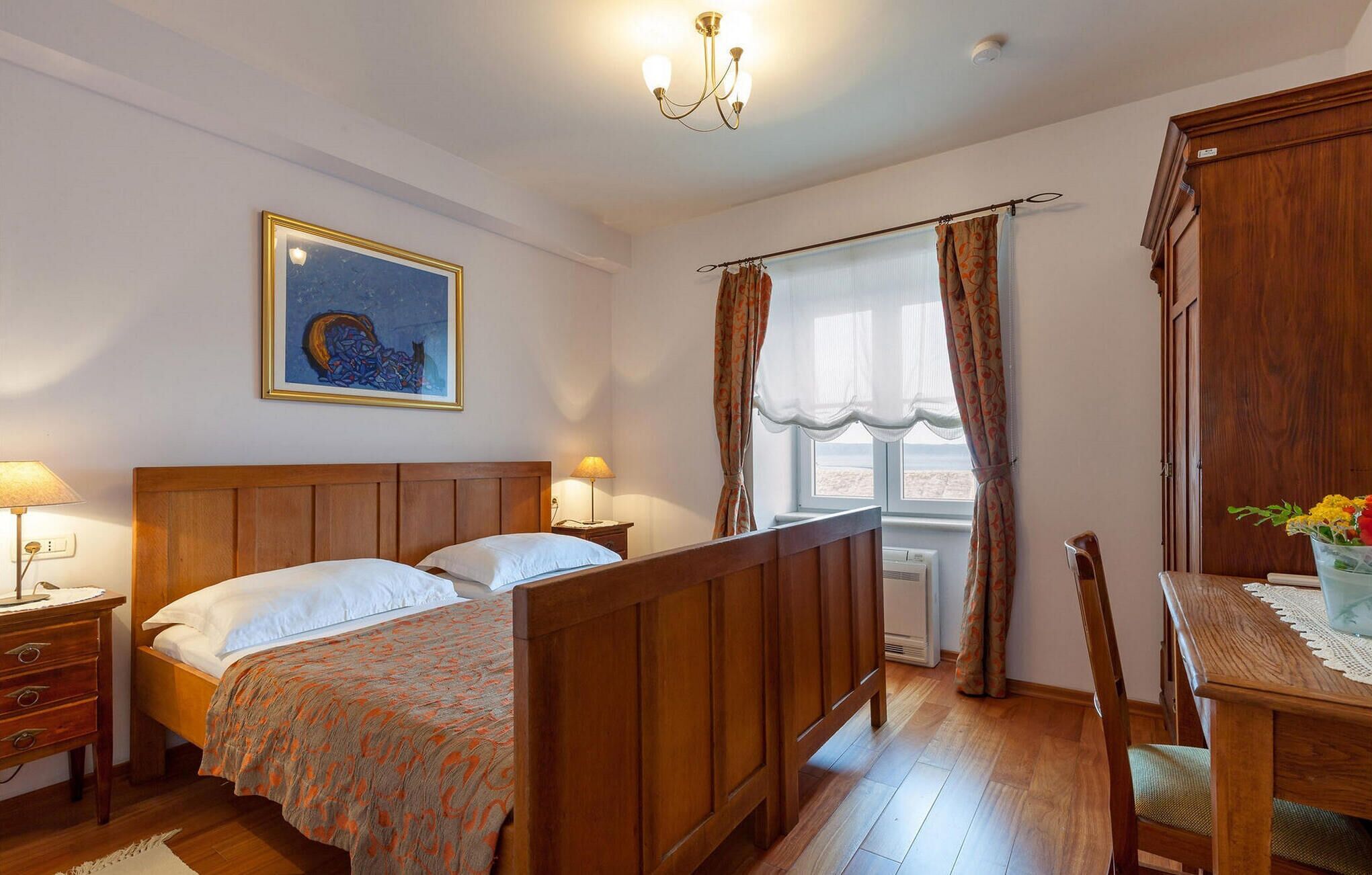 A bedroom with a wooden bed, two bedside tables with lamps, a framed artwork on the wall, a window with curtains, and a wooden wardrobe.