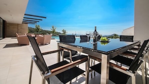 Un área de comedor exterior moderna con una mesa y sillas de vidrio, una piscina al fondo y vistas del paisaje.