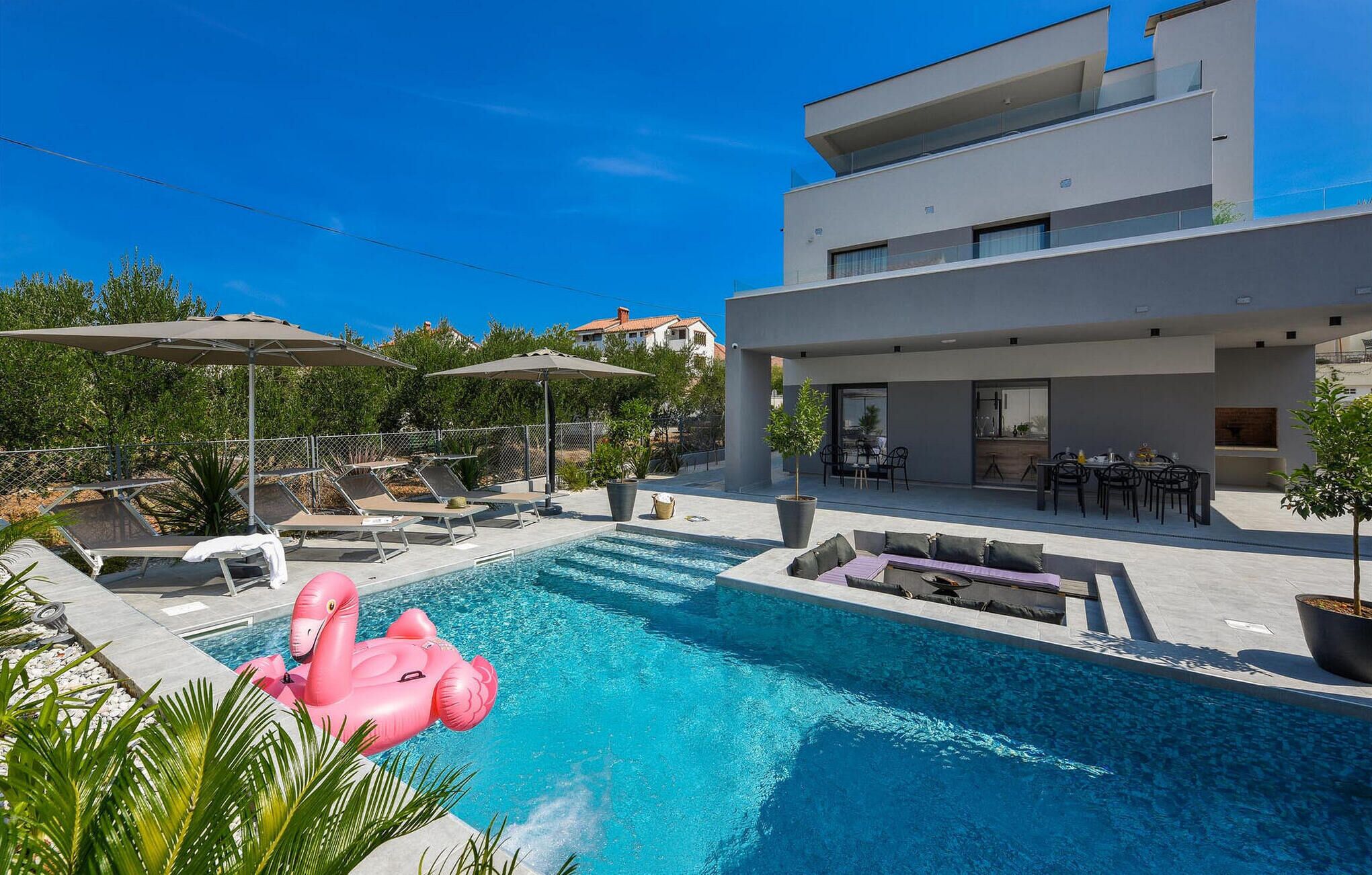 Un área de piscina moderna con sillones de descanso, un flotador con forma de flamenco rosa y una zona de comedor con asientos exteriores.