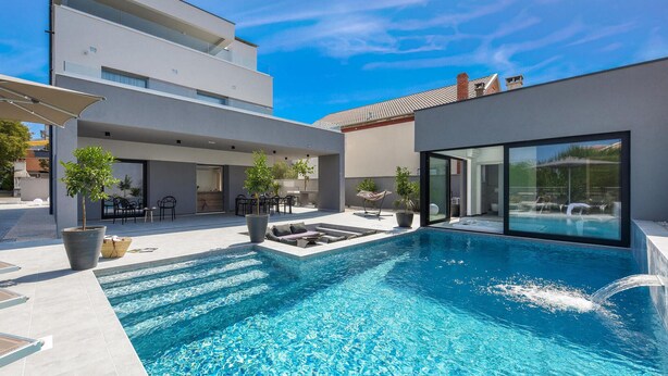 Un área de piscina exterior moderna con una piscina azul cristalina, sillones de descanso y plantas en macetas.