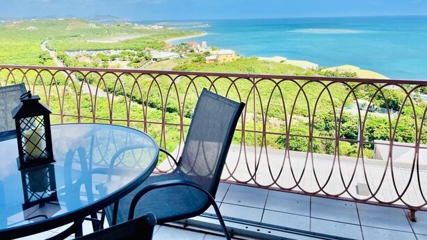 Un balcone con tavolo e sedie in vetro, affacciato su un paesaggio costiero con vista libera sul mare e colline lontane.