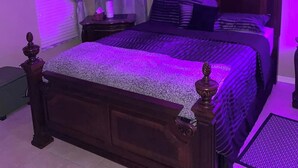 Quarto com cama de madeira, ventoinha no teto e iluminação roxa ambiente.
