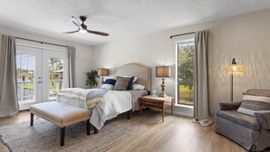 A bedroom with a bed, bedside tables, a bench, a ceiling fan, and a view through a large window.