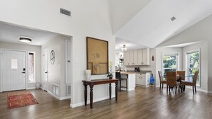 A spacious living area with hardwood flooring, a dining table with chairs, a kitchen with white cabinets, and a large window providing natural light.