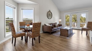 A spacious living room with a high ceiling, a dining area with wicker chairs, a brown leather sofa, and a wooden coffee table.