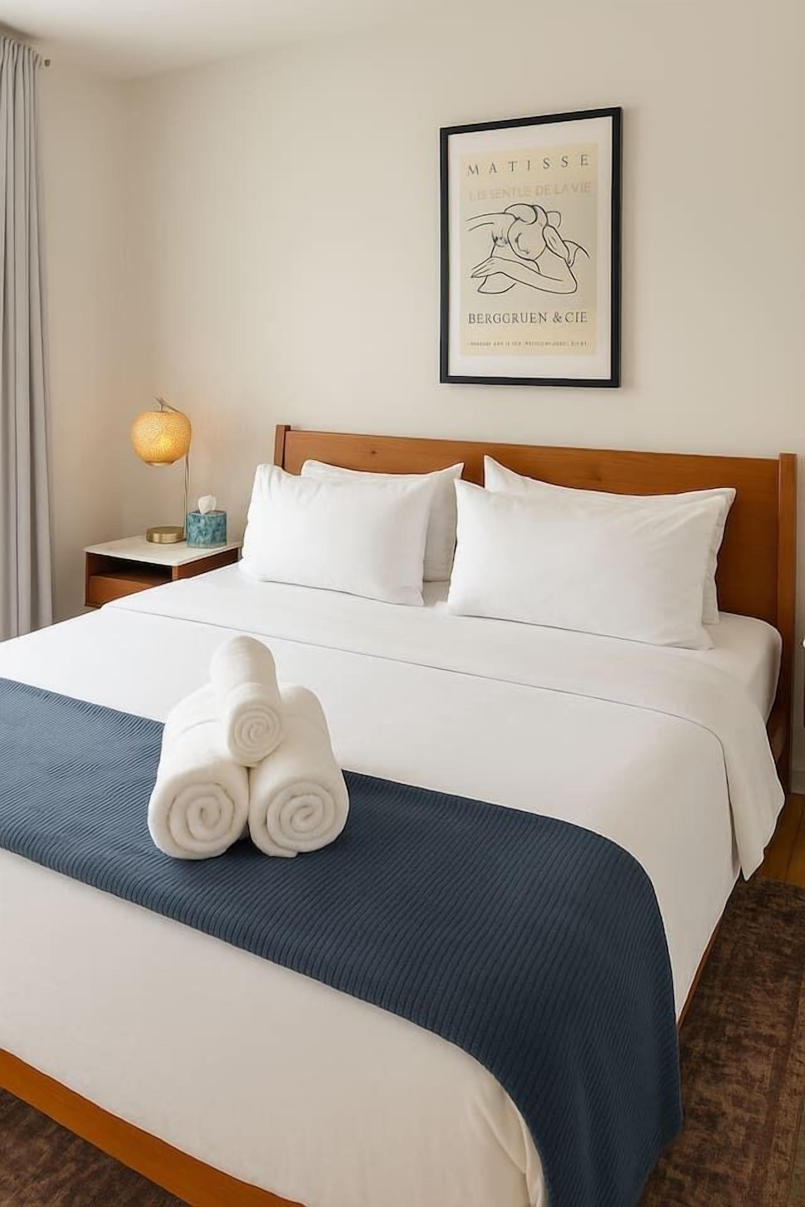 A hotel room with a neatly made bed, a framed artwork on the wall, and a bedside lamp.