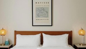 Een net opgemaakt bed met witte lakens, twee kussens en opgerolde handdoeken op een blauwe sprei. Daarboven hangt een lijst met het kunstwerk 'I Essentiel de la Vie' van Matisse.